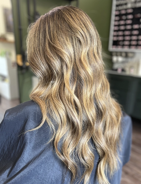 Stylish woman viewed from the back with soft, wavy blonde hair featuring caramel highlights, in a modern hair salon.