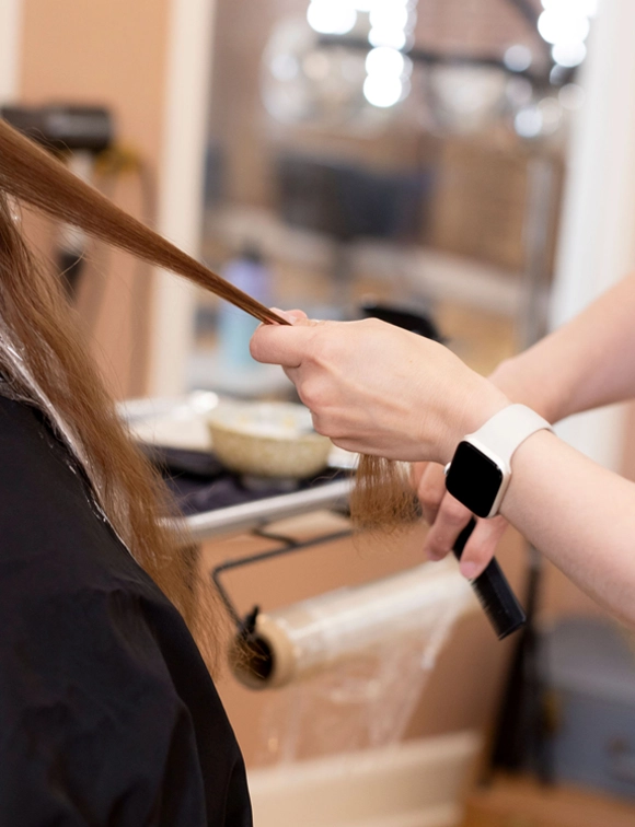 A hairstylist, wearing a smartwatch, carefully holds and trims a long strand of brunette hair in a well-equipped salon, highlighting the precision and attention to detail in the hairstyling process.