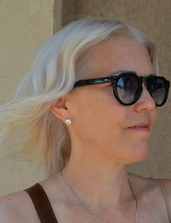 A close-up portrait of a middle-aged woman with stylish buttercream blonde hair swept to the side. She wears large dark sunglasses and diamond earrings, with a hint of a smile, portraying a casual yet elegant outdoor look.